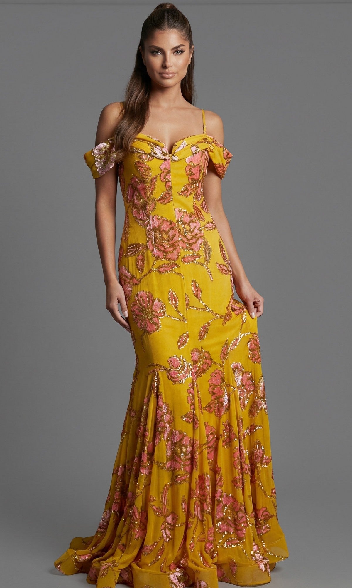 A woman wearing the Long Formal Dress: Jovani 44652—a yellow gown with pink floral embroidery—stands against a plain gray background.